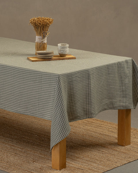 El mantel de lino menique en color verde a cuadros, cayendo sobre una mesa de madera, con un jarrón, una tabla de madera y un juego de platos para postre con pequeños cuencos de barro encima.