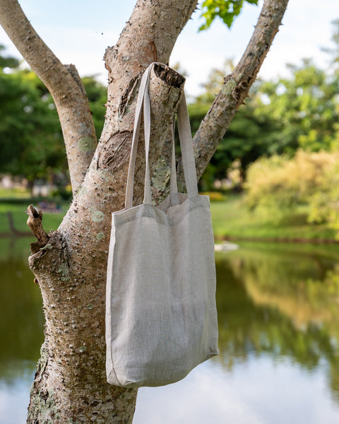Bolsa de lino color natural Menique con asas largas colgada de una rama de un árbol de tono claro. En el fondo desenfocado, hay un cuerpo de agua que refleja el cielo, rodeado de hierba verde y árboles bajo un cielo parcialmente nublado.
