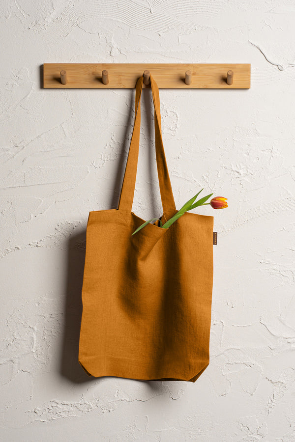 Bolsa tote duradera de lino amarillo picante Menique cuelga de un perchero de madera con cuatro ganchos montado en una pared texturizada de color blanco roto.
