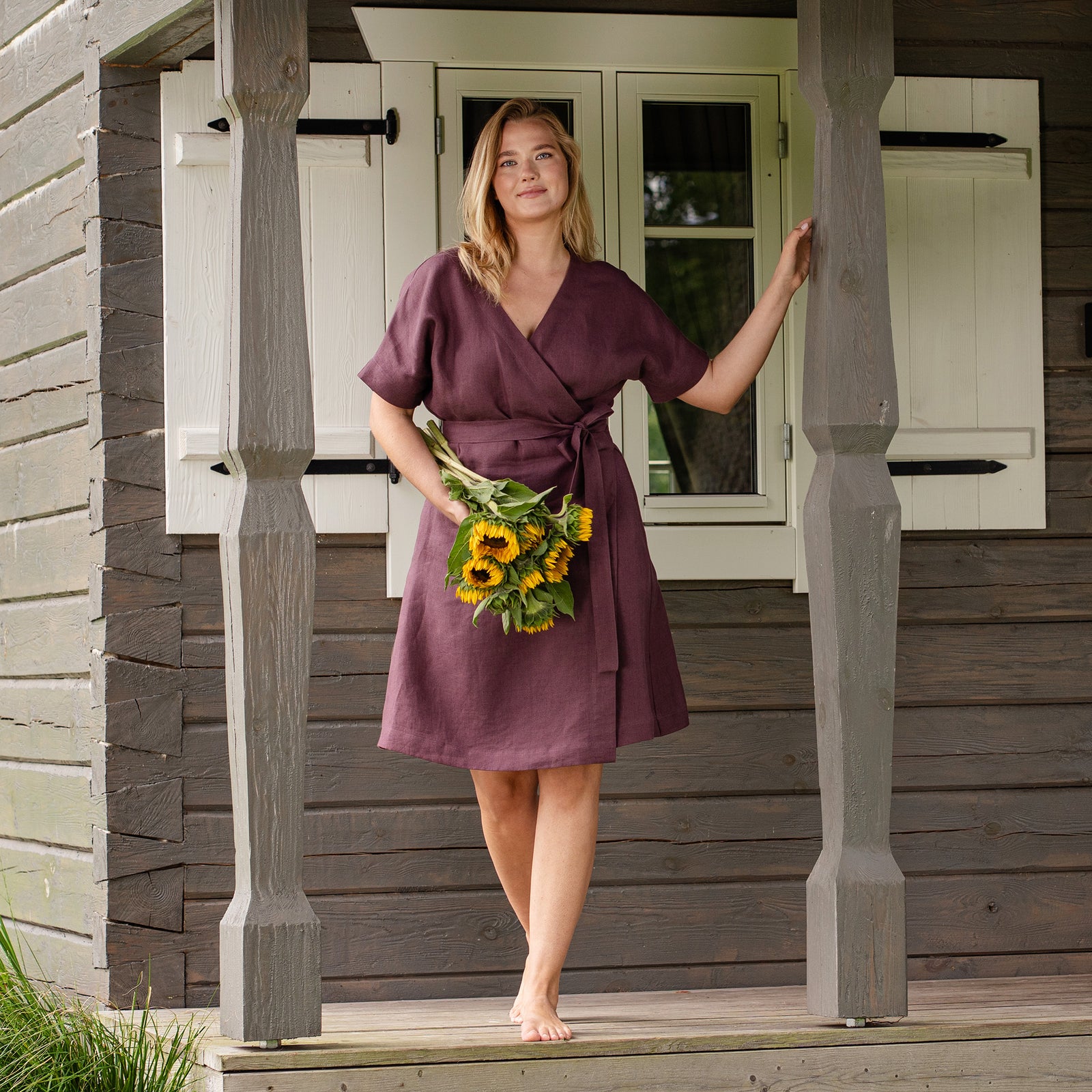 A barefoot woman poses on the wooden porch of a rustic house, resting one hand on a decorative pillar. She is wearing a knee-length, shadow purple linen wrap dress with a tie waist and holding a bouquet of fresh sunflowers.