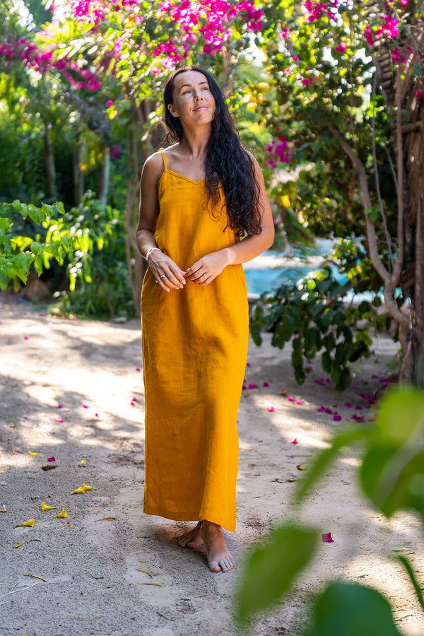 Una mujer con cabello largo, oscuro y ondulado está descalza sobre un sendero arenoso, mirando hacia arriba a la izquierda. Lleva un vestido de lino color mostaza con tirantes finos. La rodean plantas y árboles verdes y frondosos, y hay flores rosadas floreciendo al fondo. Pétalos amarillos están esparcidos sobre el camino.