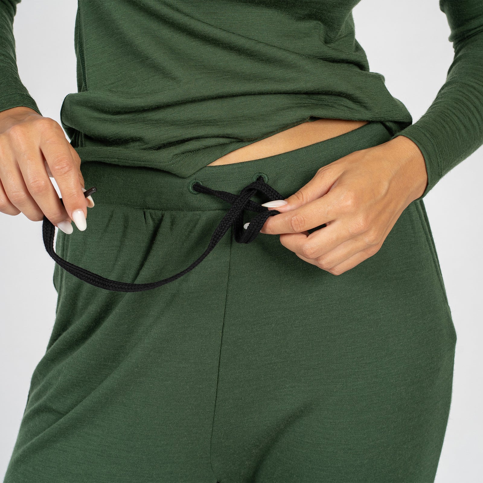 Una joven lleva puestos los pantalones deportivos de lana merina de 250 g/m² para mujer de menique y un conjunto de camiseta de manga larga. Tiene una mano detrás de la cabeza y la otra en la cadera, levantando la camiseta.