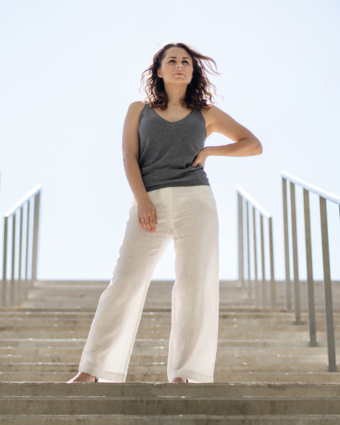 Una mujer con cabello castaño ondulado se encuentra con confianza en unas escaleras de concreto al aire libre, mirando ligeramente hacia arriba con una mano en la cadera. Lleva una camiseta sin mangas gris oscuro y pantalones blancos de lino menique, con barandillas de metal a ambos lados de las escaleras que conducen a un cielo brillante.