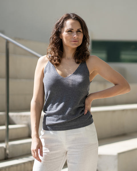 Una mujer con cabello castaño ondulado mira hacia su derecha, con la mano en la cadera, vistiendo una camiseta sin mangas gris oscuro y pantalones blancos Menique. Está de pie sobre unas escaleras de color claro con barandillas plateadas.