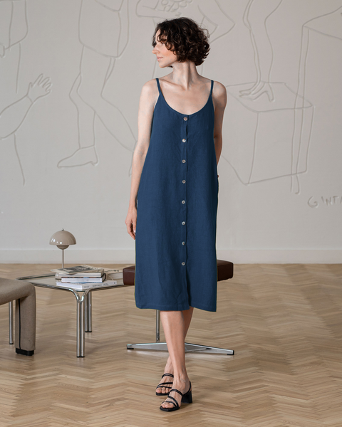 Una mujer con cabello corto, rizado y oscuro, vestida con un vestido midi azul tormenta sin mangas y con botones al frente, y sandalias negras de tacón ancho, está de pie mirando ligeramente hacia su derecha, mirando hacia la distancia. Sus manos están relajadas a los lados.