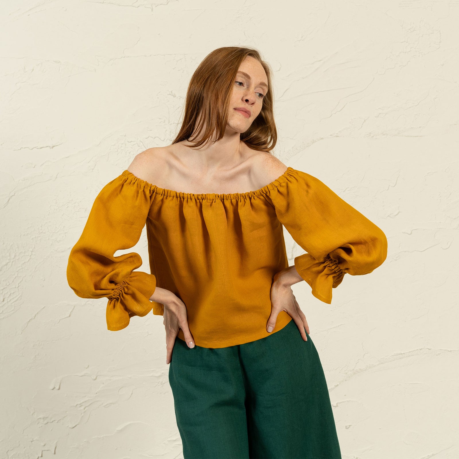 Una mujer de piel clara con cabello largo, liso y castaño rojizo está frente a una pared texturizada de color claro, mirando ligeramente hacia su izquierda. Lleva una blusa amarilla con los hombros descubiertos y mangas voluminosas recogidas en las muñecas, y pantalones verde oscuro. Tiene las manos en las caderas.