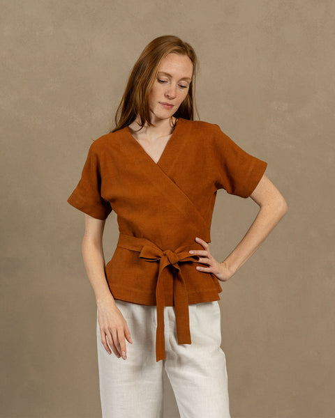 Una persona de piel clara con cabello largo y castaño rojizo, vistiendo una blusa cruzada de lino con escote en V, mangas cortas y anchas, y pantalones de color claro. Sostiene los extremos del lazo de la blusa frente a ella, formando un moño.