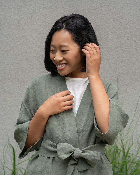 Mujer con cabello corto y oscuro que lleva una chaqueta de lino verde piedra y pantalones menique verde piedra.