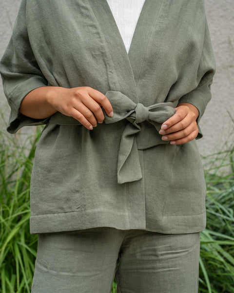 Una persona de piel clara y cabello oscuro está al aire libre, vistiendo una blusa de lino estilo kimono en verde piedra y pantalones anchos a juego. Su mano derecha está en el bolsillo de los pantalones. A su izquierda se ven hierbas altas verdes y otras plantas.