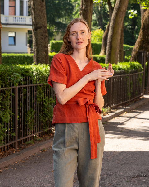 Una persona de piel clara y cabello oscuro está de perfil, mirando hacia abajo sus manos que están cruzadas a la altura del abdomen. Lleva una blusa cruzada de lino con mangas cortas y sueltas y pantalones anchos de pierna en verde piedra. Al fondo, hay una cerca de metal oscuro y un follaje verde y frondoso.