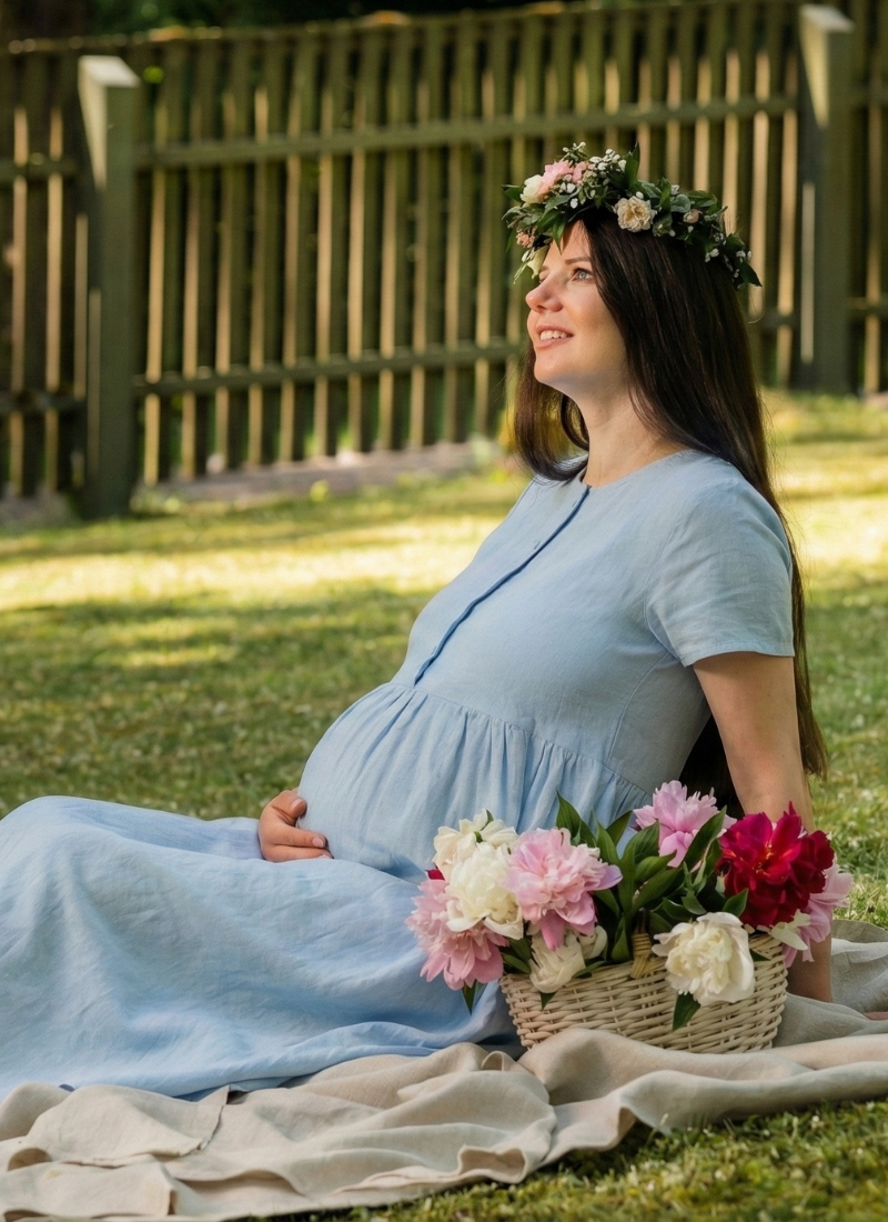 Una mujer embarazada está sentada sobre una manta en un jardín verde, vistiendo un vestido premamá de lino azul claro y una delicada corona de flores. Ella apoya suavemente su mano sobre su barriga mientras mira hacia arriba, con una cesta de flores frescas a su lado, creando una escena veraniega serena y alegre.