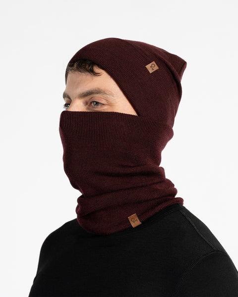 Un modelo masculino, visto de perfil, lleva un gorro de punto color cereza real y un braga de cuello a juego que le cubre la nariz y la boca. Tanto el gorro como la braga tienen un pequeño parche de cuero marrón con el logo de Menique. Tiene barba corta y cabello oscuro. El fondo es blanco sólido.
