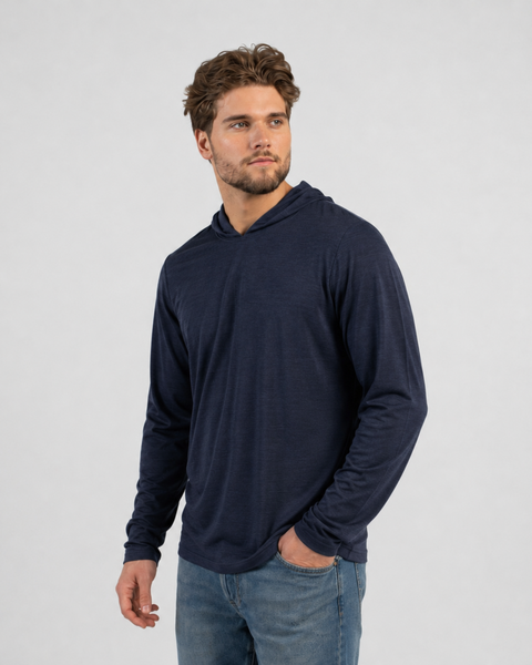 A man with wavy brown hair and a beard posing against a plain light background, wearing a lightweight, long-sleeve navy blue hoodie with the hood down and blue jeans, with his left hand in his pocket.