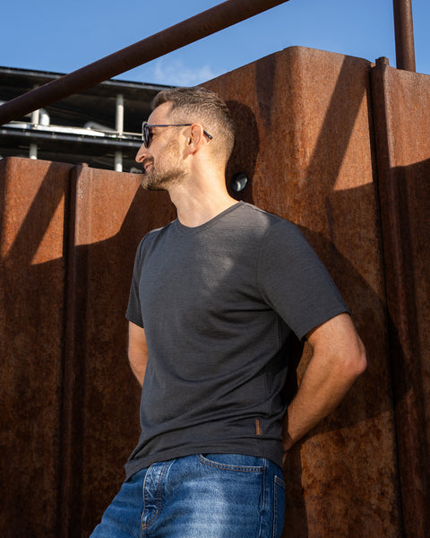 A man wearing a charcoal grey short-sleeved t-shirt, blue jeans, and sunglasses leans back against a large, rusted steel wall. His hands are resting behind his back, and he is looking off to the side under a blue sky. A small brown logo tag is visible on the lower hem of the shirt.