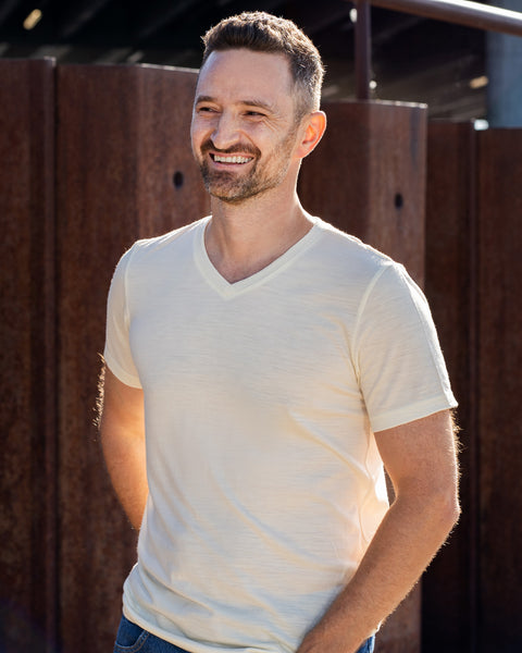 A man wearing a short-sleeved, natural off-white v-neck t-shirt and blue jeans stands outdoors with his hands behind his back. He is smiling warmly and looking to the left, framed against large, vertical rusted steel beams.