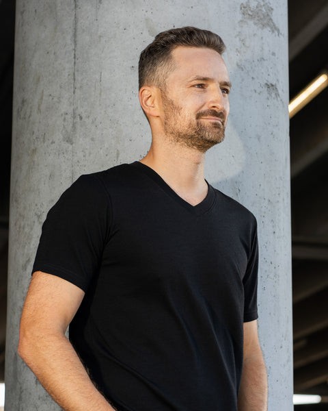 A man standing outdoors next to a large concrete pillar, looking off to the side with a slight smile. He is wearing a short-sleeve black V-neck t-shirt.