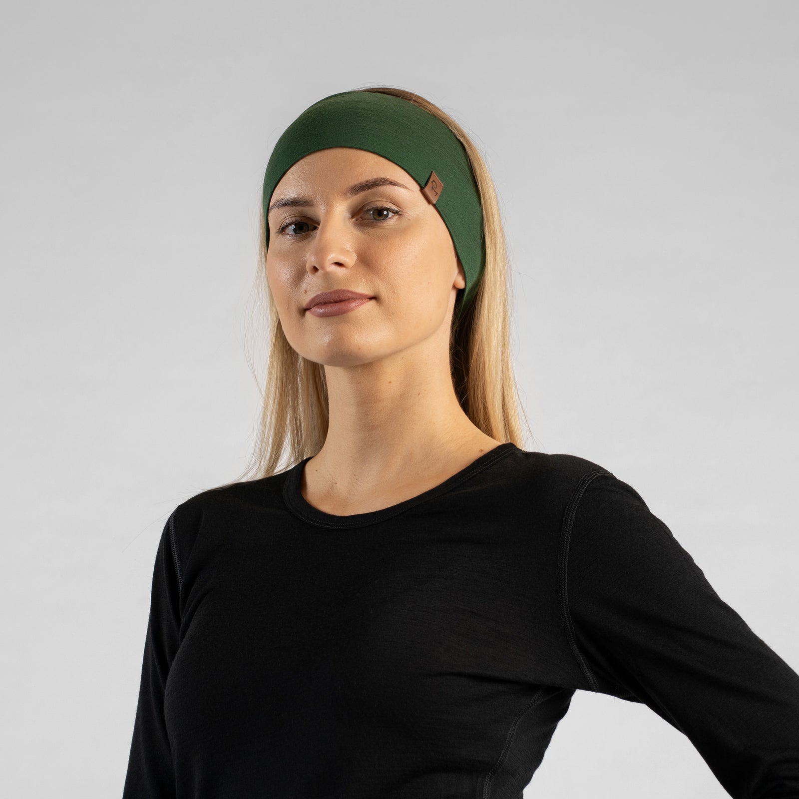 La imagen muestra a una mujer sentada en un bosque, vistiendo la cinta para la cabeza unisex menique de merino en verde oscuro. También lleva una camiseta sin mangas de merino negra. Ella mira hacia un lado.