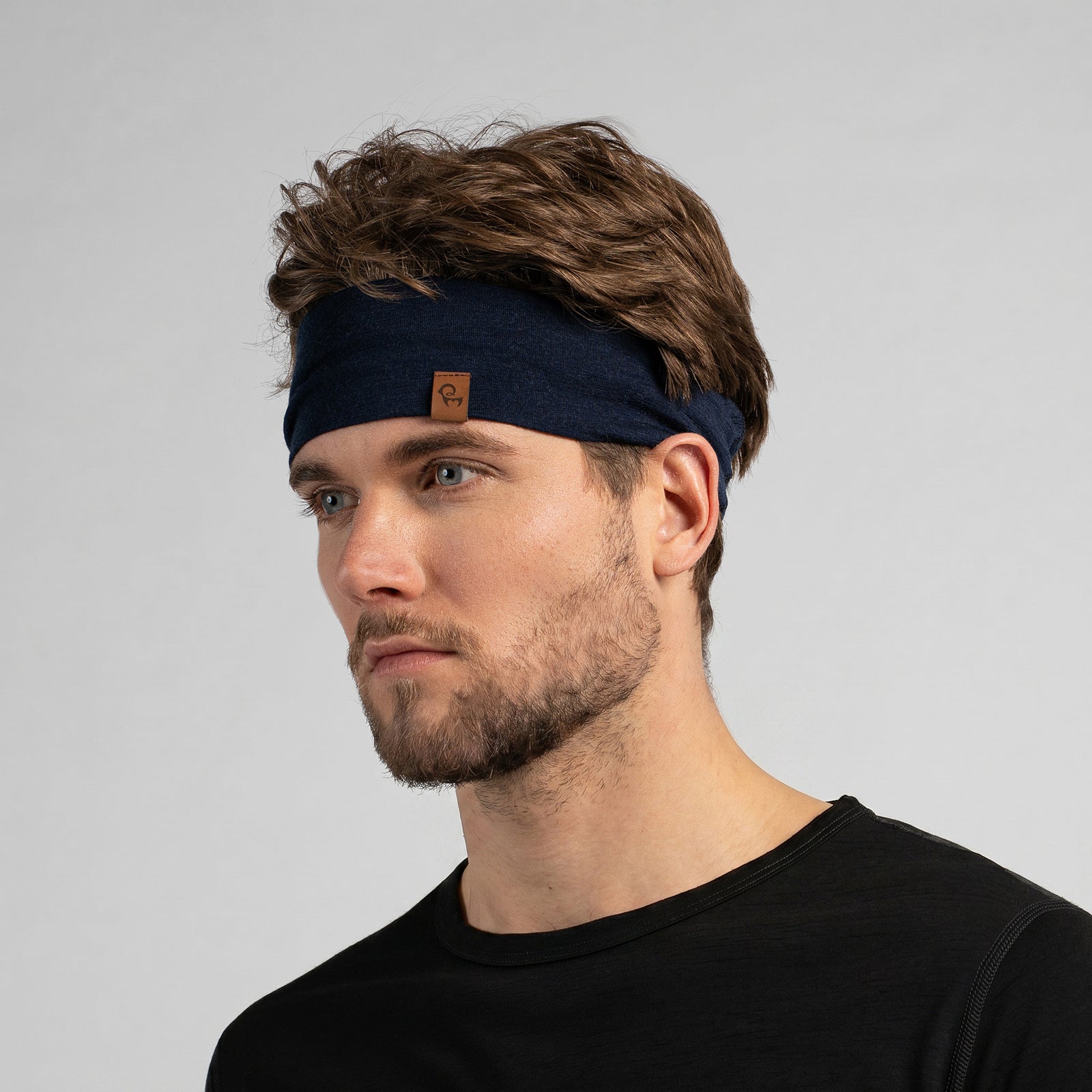 A dark navy blue, finely knitted headband displayed in a three-dimensional, rounded shape against a pure white background. A small, square brown leather logo patch is centered near the bottom front edge.