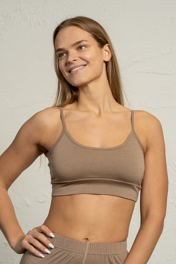 Una mujer que lleva un bralette de merino beige sonríe suavemente mientras posa con una mano en la cadera. El bralette tiene tirantes delgados ajustables y un diseño liso y ceñido. Ella está de pie frente a un fondo claro con textura.