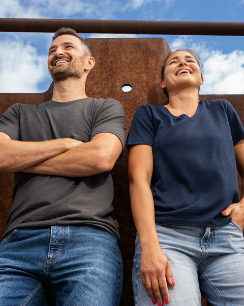 A low-angle shot of a smiling man and a laughing woman leaning back against a large, rusted metal wall under a blue sky with clouds. The man is on the left with his arms crossed, wearing a dark grey short-sleeve t-shirt and blue jeans. The woman is on the right with one hand resting in her pocket, wearing a navy blue short-sleeve v-neck t-shirt and light blue jeans.