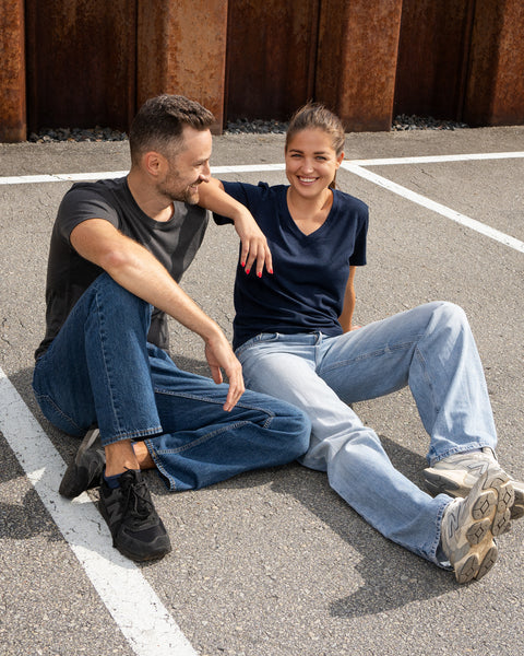 A smiling man and woman are sitting comfortably on an asphalt surface marked with white lines, looking happily at each other. The man is on the left wearing a dark grey t-shirt, blue jeans, and black sneakers. The woman leans slightly toward him with one arm resting behind his back, wearing a navy blue v-neck t-shirt, loose light blue jeans, and grey sneakers.