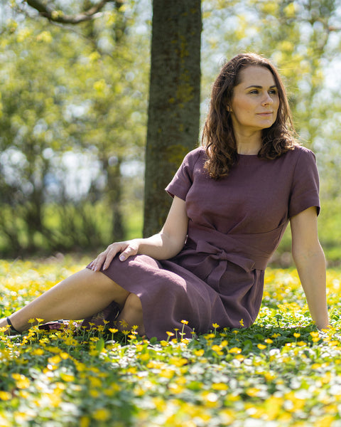 Una mujer de cabello castaño, vestida con un vestido morado de manga corta, está sentada al aire libre sobre la hierba entre pequeñas flores amarillas.