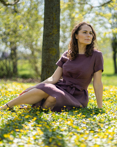Una mujer de cabello castaño, vestida con un vestido morado de manga corta, está sentada al aire libre sobre la hierba entre pequeñas flores amarillas.