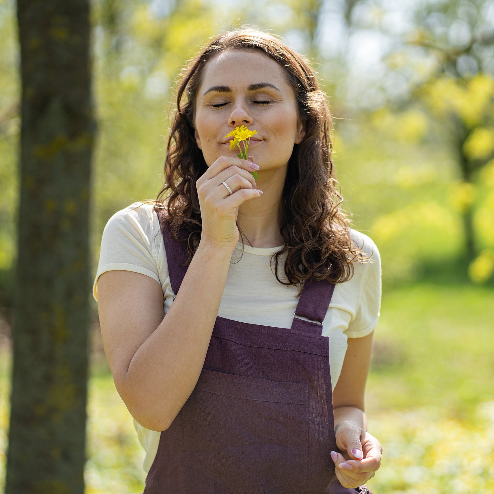 Una mujer con cabello castaño, vestida con un mono de color púrpura oscuro sobre una camiseta de tono claro, está de pie al aire libre entre dos troncos de árbol sobre un suelo cubierto de hierba.