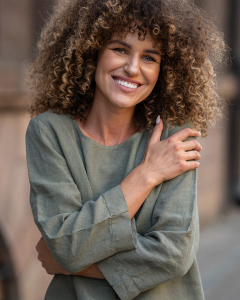 Mujer con cabello rizado que lleva una blusa verde de lino, sonriendo al aire libre.