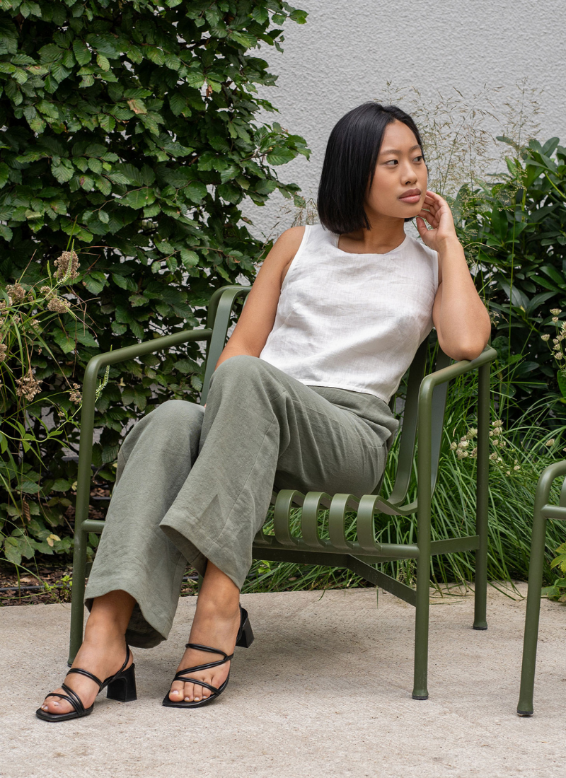 Una mujer de cabello corto y negro está sentada en una silla metálica verde en un jardín, vistiendo una blusa sin mangas de lino blanco y pantalones de lino verde oliva de corte holgado. Mira hacia un lado con una expresión serena, rodeada de abundante vegetación y plantas, creando un ambiente tranquilo al aire libre.