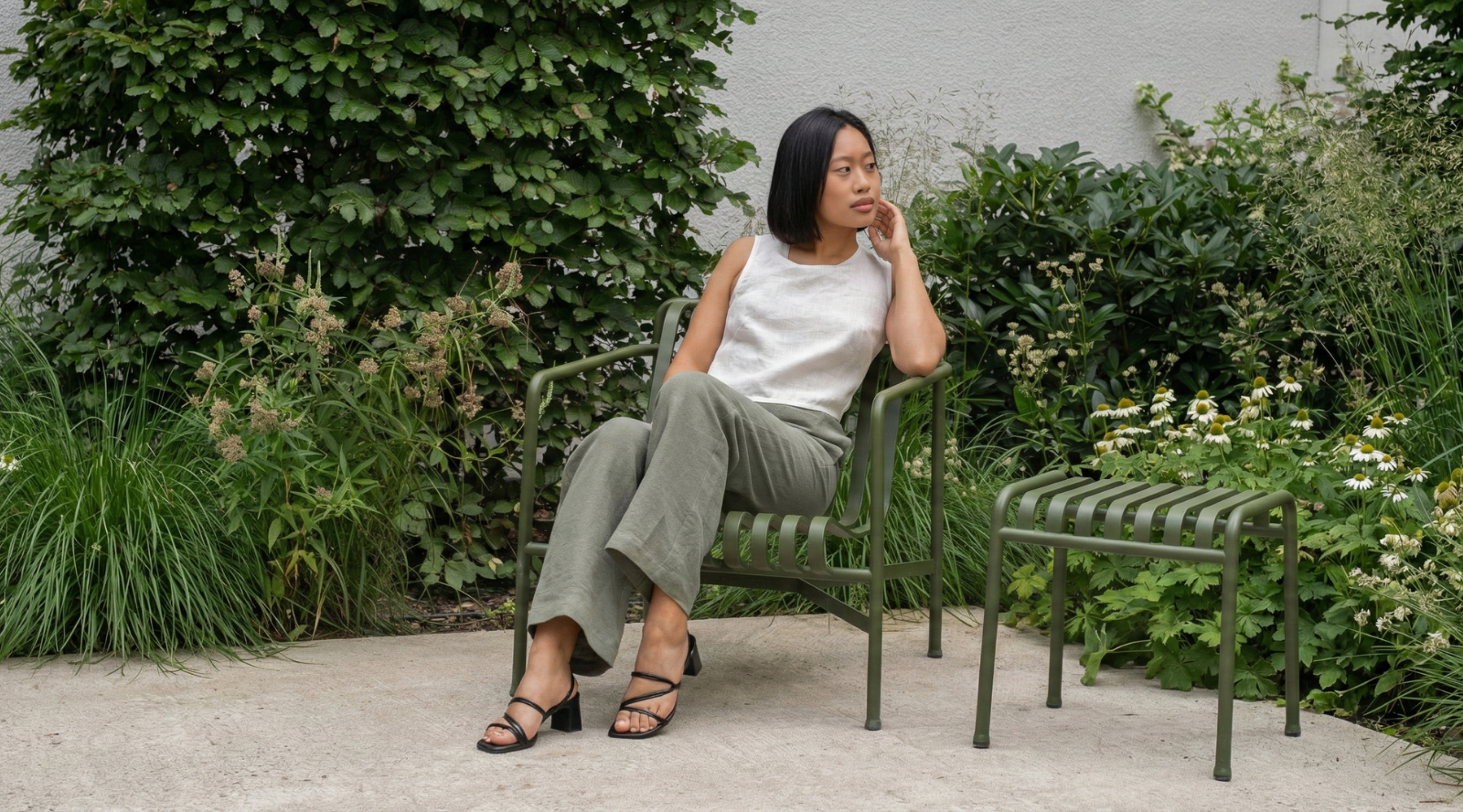 Una mujer de cabello corto y negro está sentada en una silla metálica verde en un jardín, vistiendo una blusa sin mangas de lino blanco y pantalones de lino verde oliva de corte holgado. Mira hacia un lado con una expresión serena, rodeada de abundante vegetación y plantas, creando un ambiente tranquilo al aire libre.