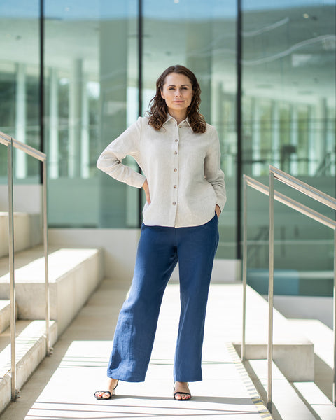 Una mujer de cabello castaño, vestida con una camisa de lino natural con botones al frente y pantalones blancos de pierna ancha, está de pie al aire libre frente a una estructura moderna de hormigón.