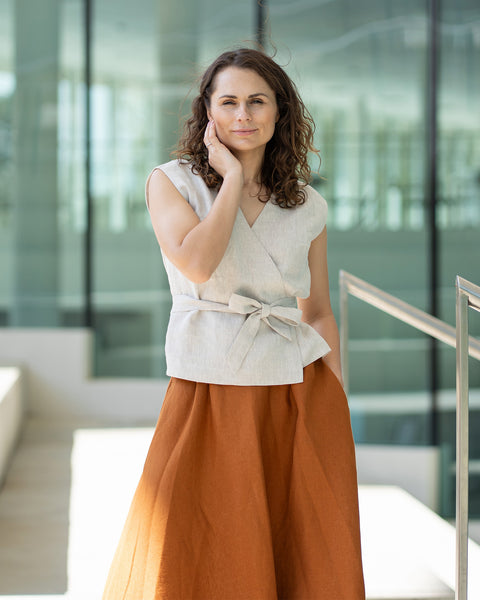 Una mujer de cabello castaño, vestida con una blusa cruzada de color beige claro y una falda larga color óxido, posa en el interior frente a grandes ventanales.