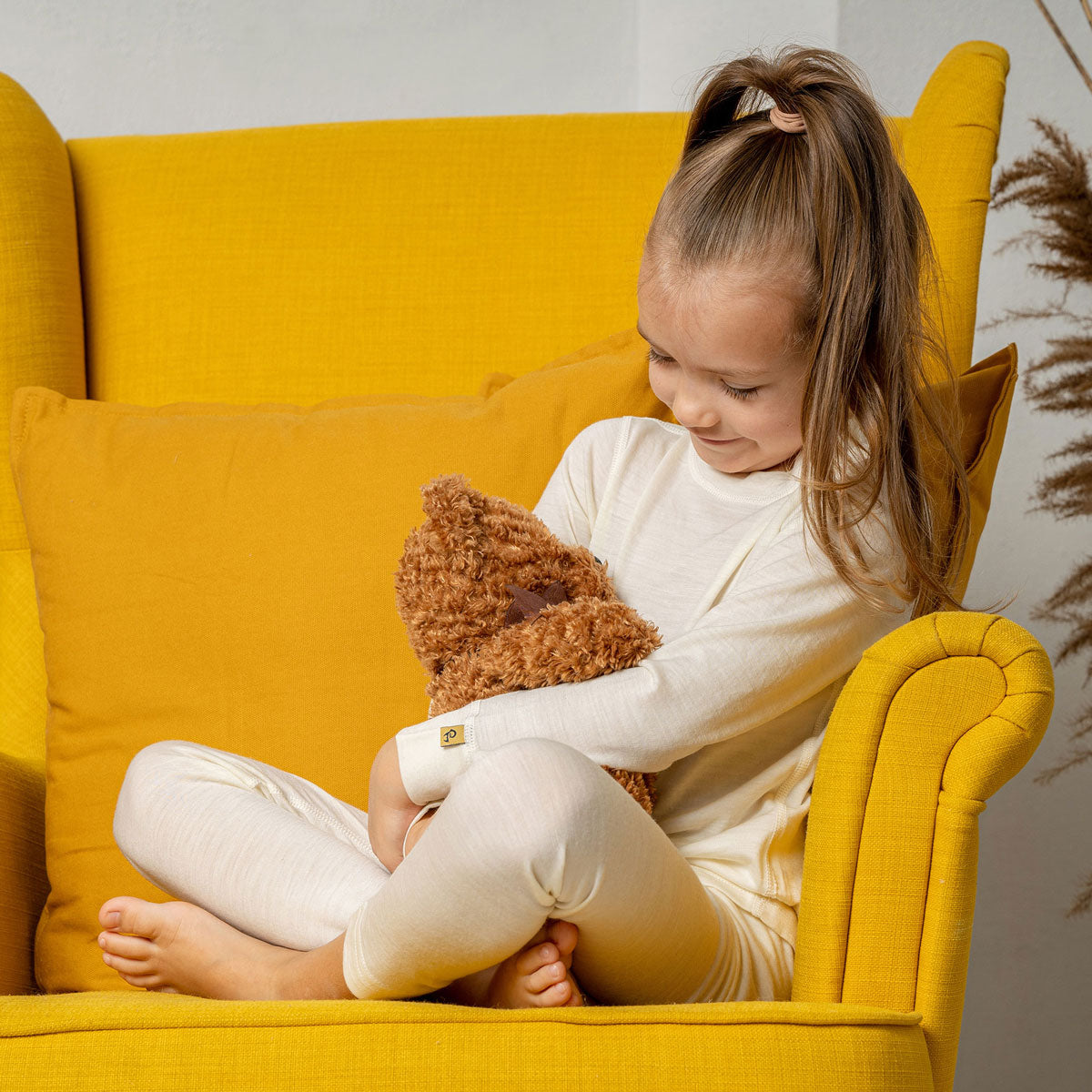 Una niña sentada en un sillón amarillo, con las piernas cruzadas, vistiendo la camiseta de manga larga menique de merino 160gsm en color natural.