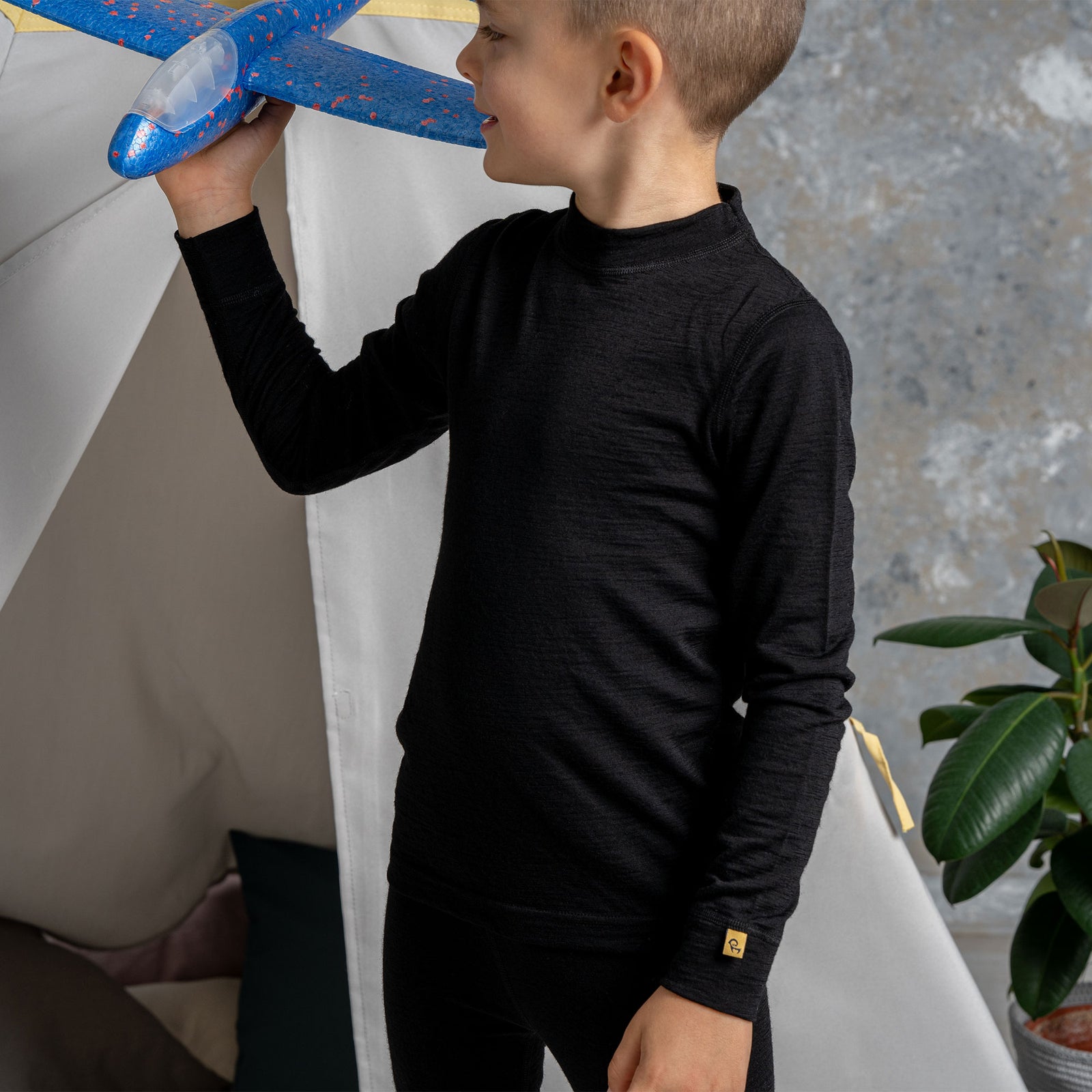 Un niño pequeño con cabello rubio y ojos azules, vestido con una camiseta negra de manga larga y pantalones vaqueros azules, se está colgando de una barra metálica en una estructura de parque infantil. Mira directamente a la cámara con una expresión feliz, con la boca abierta y la lengua ligeramente afuera.