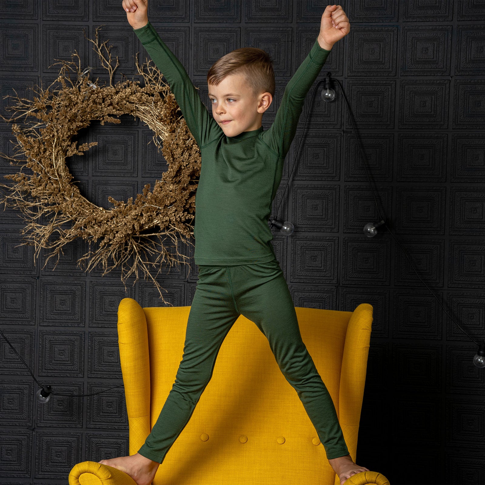 Un niño sentado en una silla, con una pierna apoyada sobre ella, vistiendo los Pantalones Merino 160 para niños de menique en color Verde Oscuro. Apoya la cabeza sobre la rodilla de la pierna levantada.
