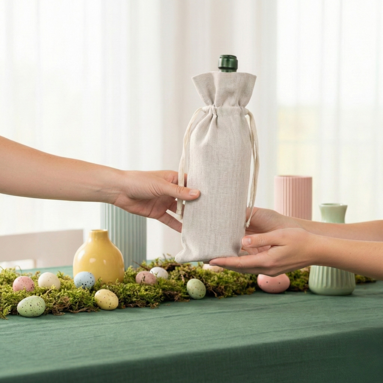 Tres bolsas de regalo para botellas de vino de lino en tonos rojo, verde y natural, exhibidas sobre una mesa primaveral con flores, velas y decoración en tonos pastel.