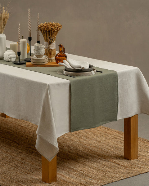 El mantel de lino menique en color blanco puro, cayendo sobre una mesa de madera, con un jarrón y un juego de taza y plato de barro encima.