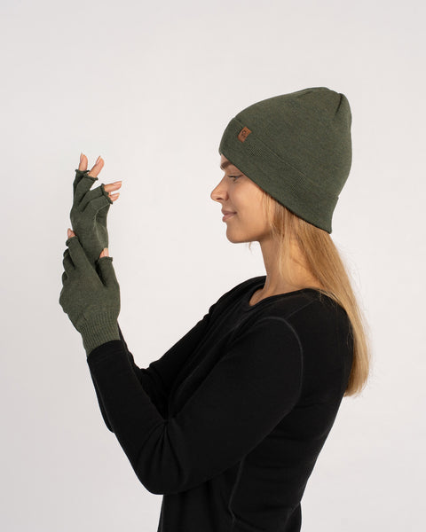 Vista lateral de una mujer que lleva un gorro de punto verde oscuro y guantes sin dedos a juego, inspeccionando sus manos levantadas.