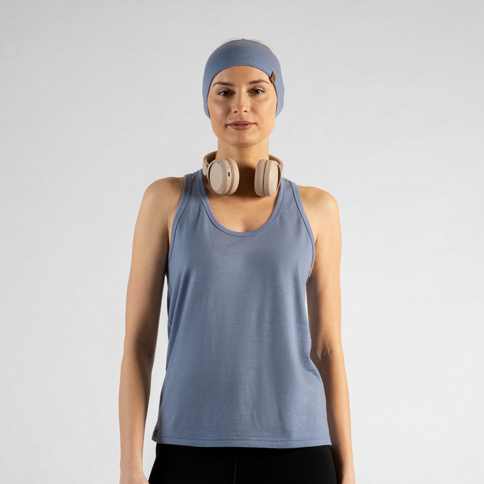 A woman sitting on a white studio floor, viewed from behind. She is wearing a dusty blue racerback tank top, black leggings, and a matching dusty blue headband. Her blonde hair is pulled into a low ponytail, and she has both hands raised behind her head as she adjusts the headband.