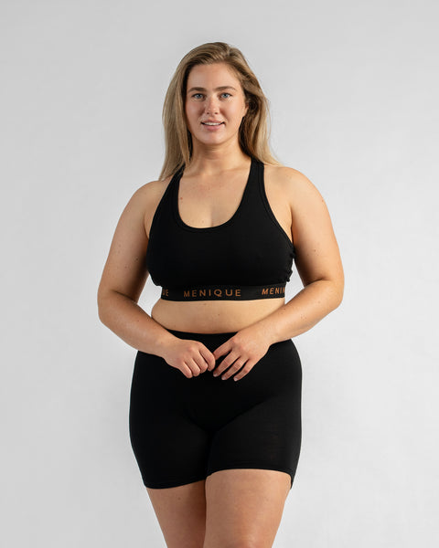 Front view of a model with her hands clasped in front of her, wearing a black sports bra and black biker shorts, both featuring a black elastic band with text.