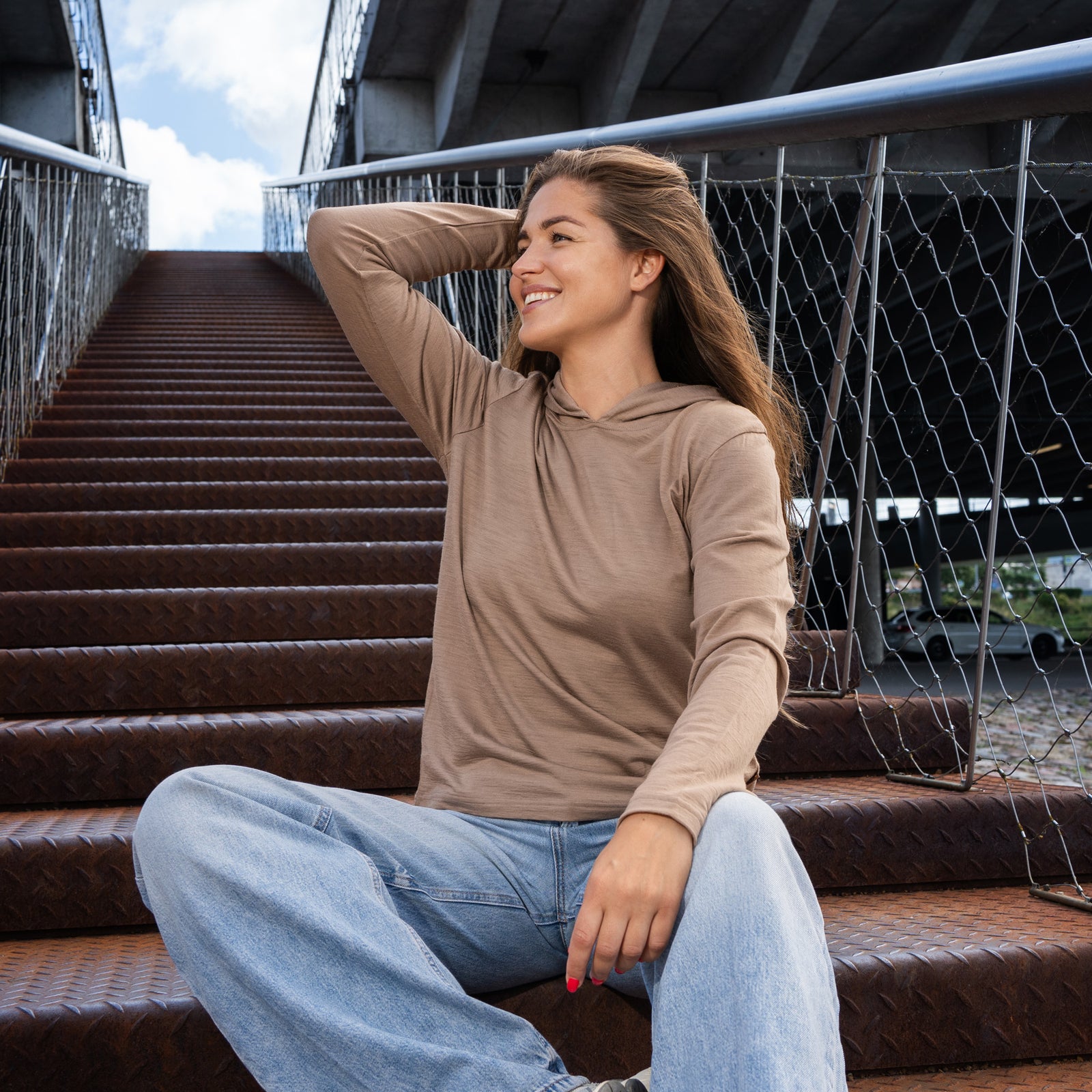 A side-profile studio portrait of a woman looking back over her shoulder at the camera against a plain light grey background. She is wearing a lightweight beige long-sleeved hoodie with the hood pulled up over her head and light wash denim jeans.