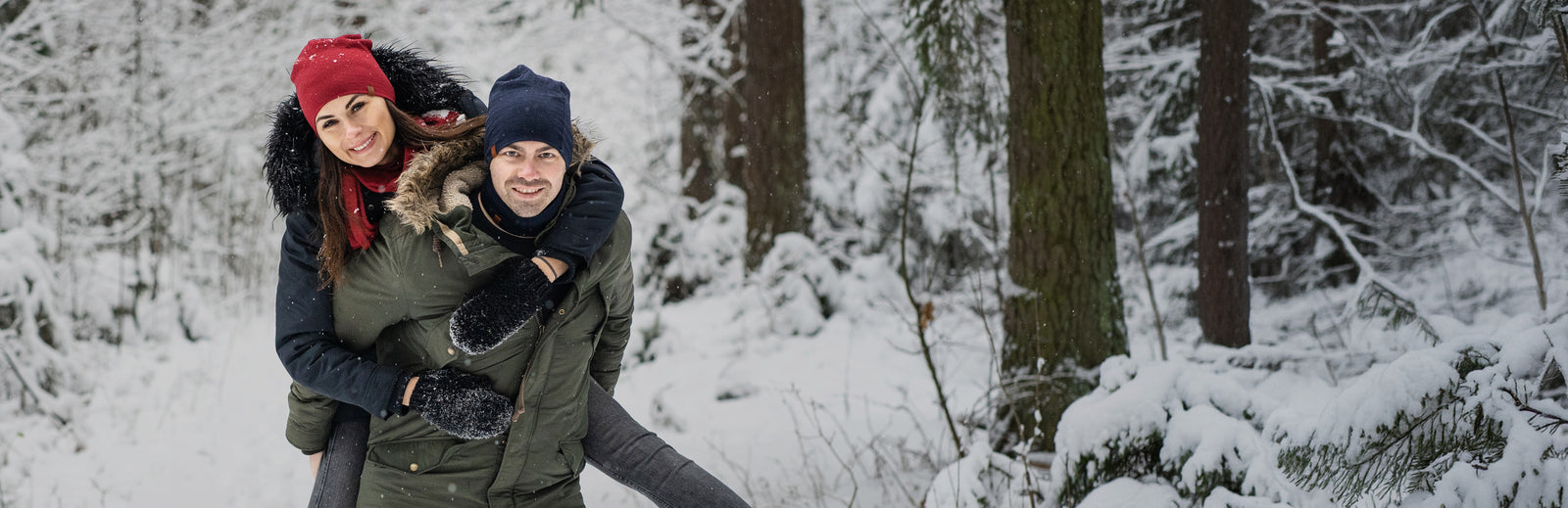 Pareja disfrutando del invierno al aire libre, mujer con gorro rojo abrazando a un hombre por detrás en un bosque nevado