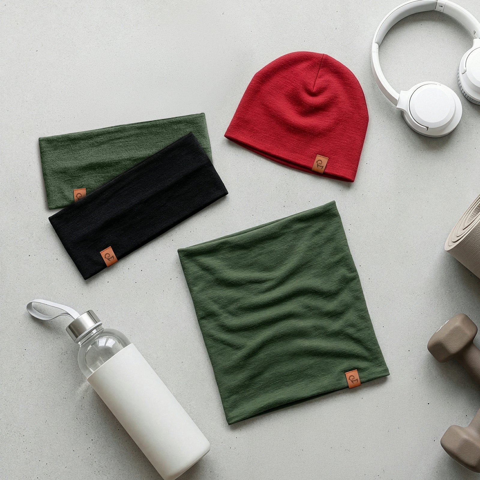 Disposición plana de cuellos tubulares de merino y un gorro rojo sobre una superficie clara con una botella de agua y auriculares.