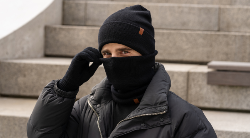 A person dressed in all-black winter accessories, including a knit beanie, neck gaiter, and gloves, poses in front of blurred stone steps while wearing a dark jacket.
