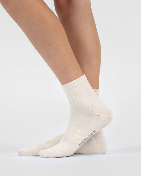 A side-profile view of a person's feet wearing natural off-white crew socks, revealing the word "MENIQUE" printed on the sole of the right foot.