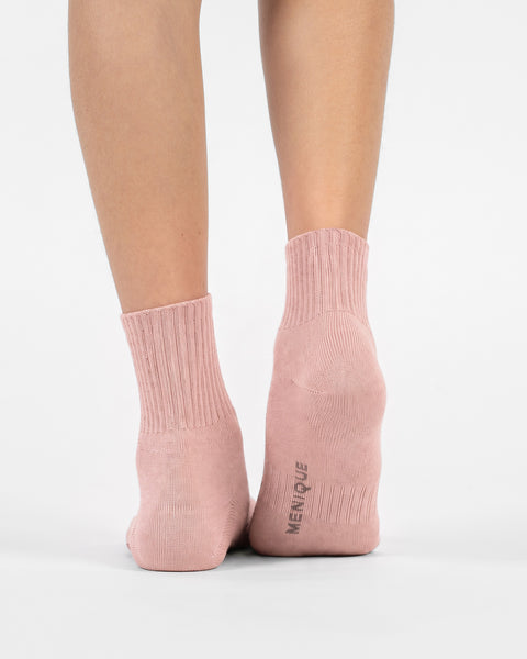 A back view of a person's lower legs and feet wearing light pink crew socks, with the right heel slightly lifted to show the "MENIQUE" logo on the sole.