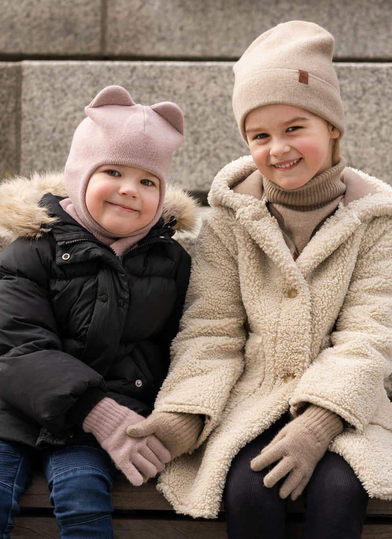 Two young children sit outdoors against a backdrop of light gray stone steps, bundled up in warm winter clothing. The child on the left is wearing a black puffy coat with a faux-fur hood and a dusty pink knit balaclava that features small animal ears on top, while the child on the right smiles brightly wearing a cream-colored textured sherpa coat, a beige ribbed turtleneck, and a matching beige beanie.