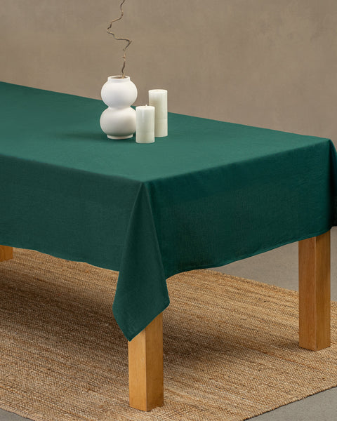 El mantel de lino menique en color verde oscuro, cayendo sobre una mesa de madera, con un jarrón blanco moderno encima y dos velas blancas de diferente altura a los lados.