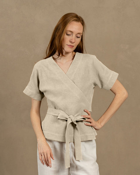 Una persona de piel clara con cabello largo y castaño rojizo, vistiendo una blusa cruzada de lino natural con escote en V, mangas cortas y anchas, y pantalones de color claro. Sostiene los extremos del lazo de la blusa frente a ella, formando un moño.